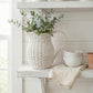 Wicker pitcher with greenery on a white wooden shelf with bowls and a towel.