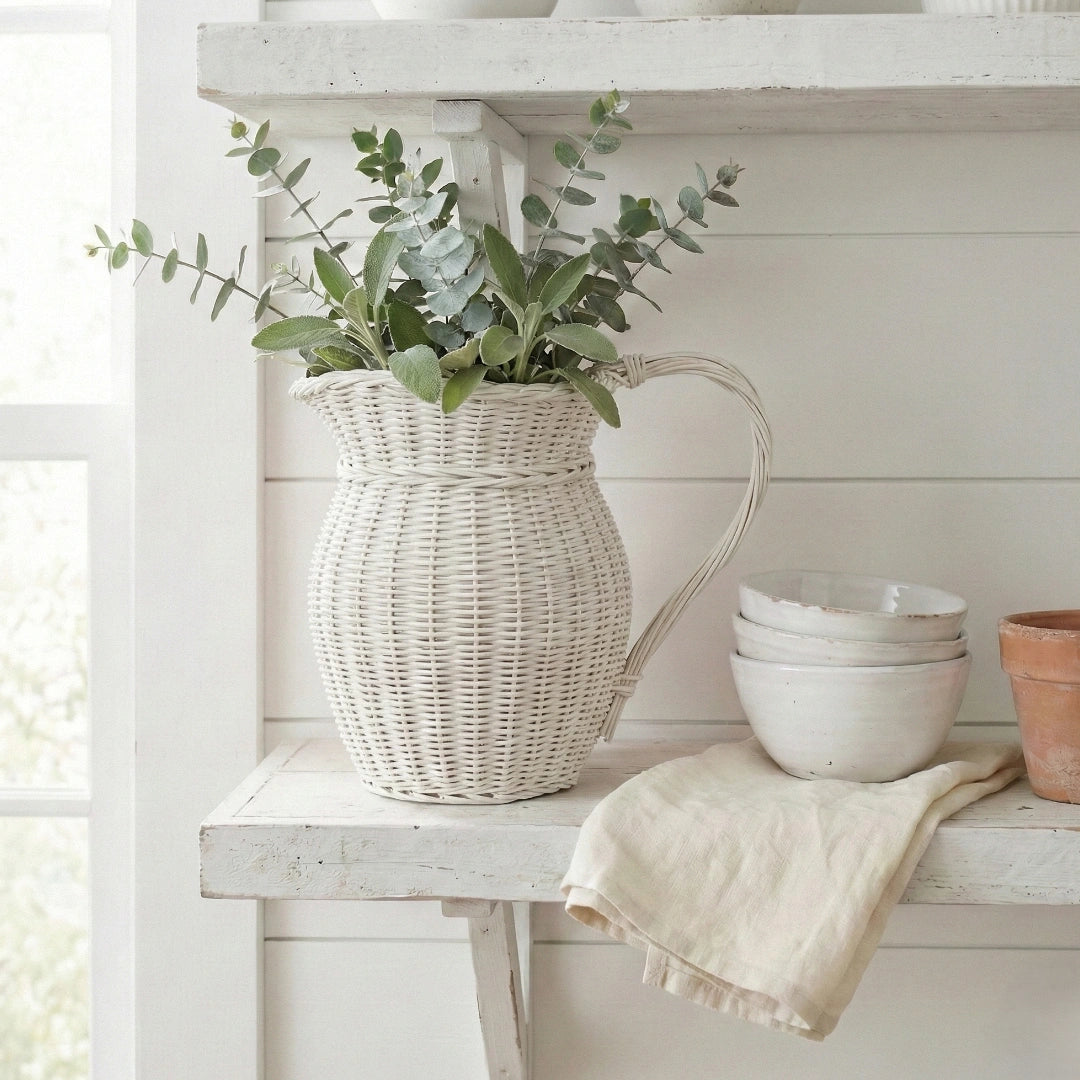 Wicker pitcher with greenery on a white wooden shelf with bowls and a towel.
