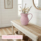 Pink woven pitcher with flowers on a wooden bench in a home interior setting.
