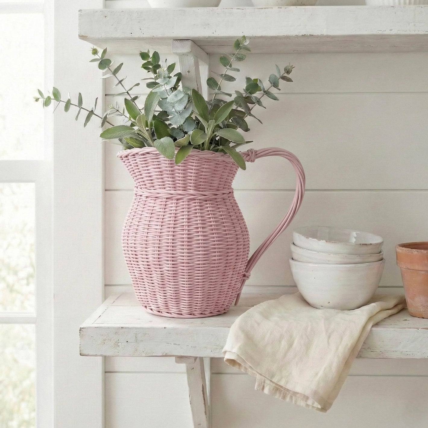 Pink wicker pitcher with greenery on a white wooden shelf