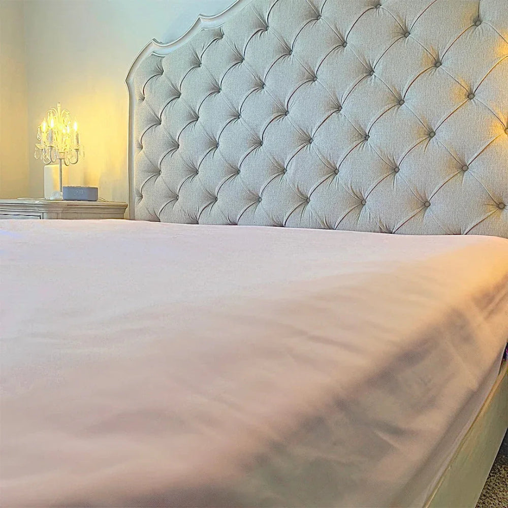 Close-up of a fitted mattress sheet on a bed with a grey tufted headboard and a lit bedside lamp.