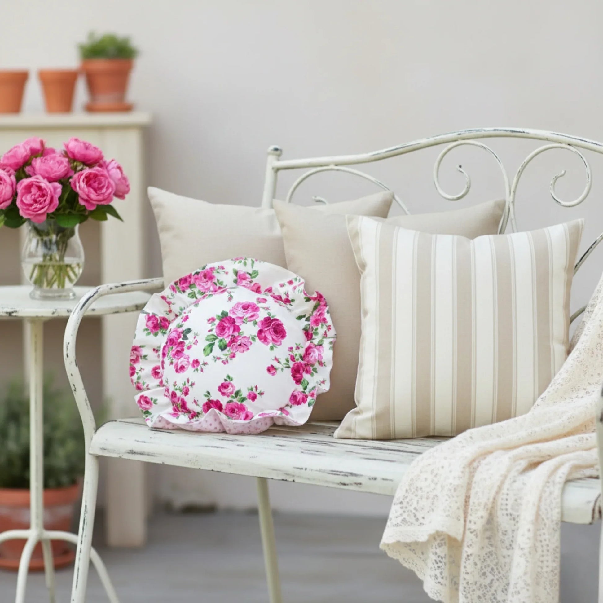 Antique white circular floral pillow on a metallic bench next to metallic tale and transparent vase with flowers on it.