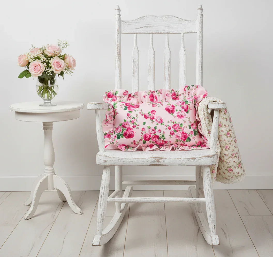 White rocking chair with pale pink rectangular floral cushion and blanket, next to a small round table with flowers, on a light wooden floor.