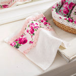 A white face towel with a floral fleur print and lace detailing, displayed on a white surface next to a wicker basket with a pink and white floral design.