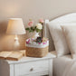 Napkin with floral pattern on a basket on a nightstand next to a bed with pillows and a lamp.