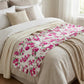 White ruffled bed runner with a pink rose floral pattern draped over neutral-toned pillows and textured blankets.