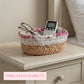 A fleur oval halfa woven basket with a pink rose fabric liner and lace trim, holding eyeglasses and a remote control on a vintage white nightstand.
