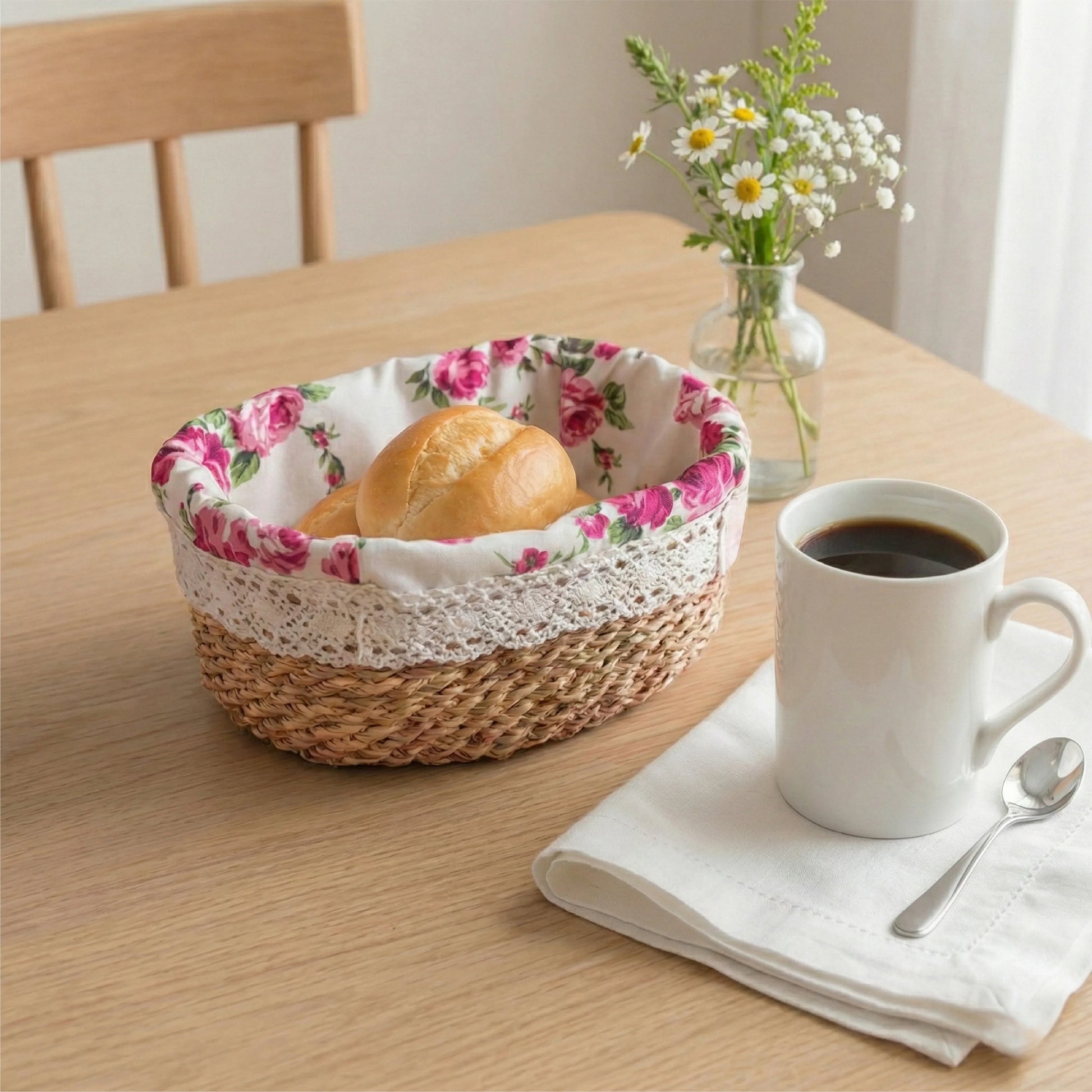 A fleur oval halfa woven basket with a signature pink rose liner and white lace trim, serving bread rolls on a wooden dining table next to a cup of coffee and a vase of daisies.