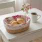 A fleur oval halfa woven basket with a signature pink rose fabric liner and white lace trim, serving two crusty bread rolls on a distressed white wooden table beside a cup of coffee and a vase of pink roses.