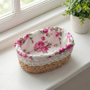 A small oval halfa grass basket with a pink rose fabric liner and white lace trim, sitting on a sunlit white windowsill next to a potted plant.