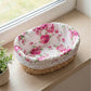 A high-angle view of a Fleur oval halfa basket with a pink rose fabric liner and white lace trim, displayed on a light wood windowsill beside a small green plant.