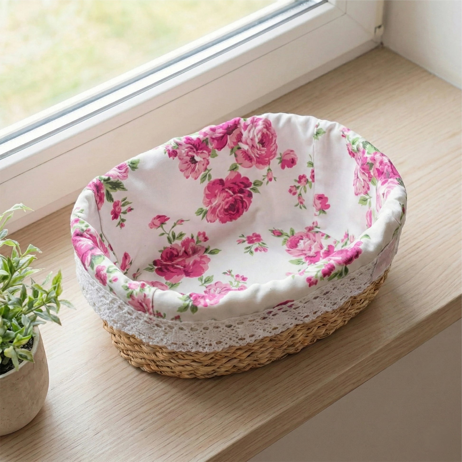 A high-angle view of a Fleur oval halfa basket with a pink rose fabric liner and white lace trim, displayed on a light wood windowsill beside a small green plant.