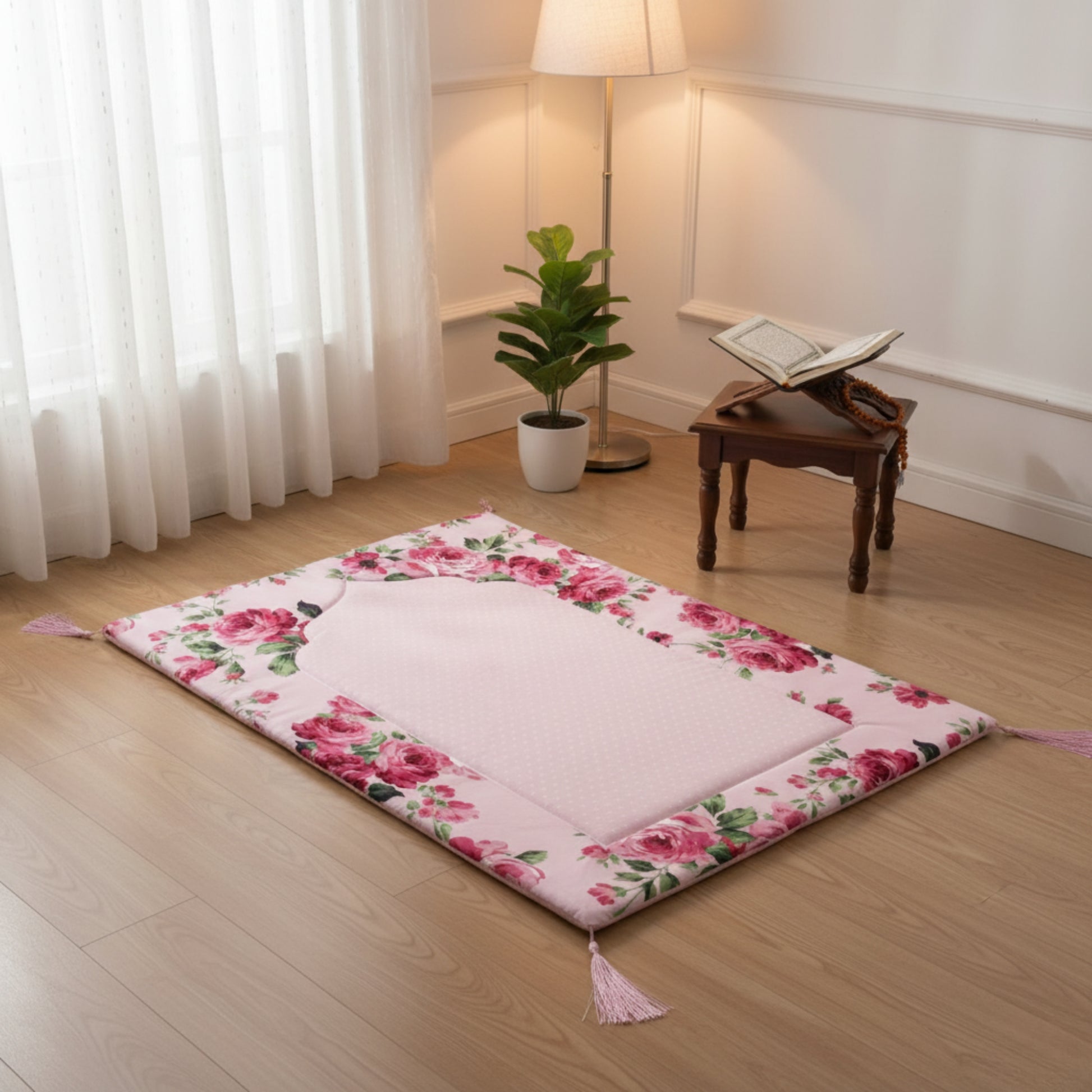 Floral-patterned pink prayer mat on wooden floor with wooden stand, Quran, plant, and lamp in the background.