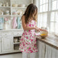 Person whisking eggs in a pink floral apron by a wooden counter in a white kitchen with open shelving.