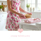 Person in a pink floral apron holding a steaming dish above a pink heart tray and the text "COOK WITH LOVE."