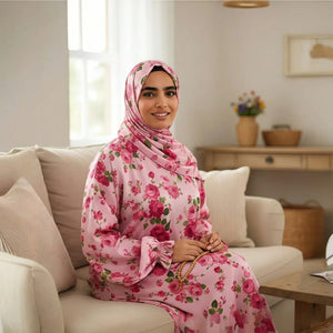 A woman wearing a pink floral hijab and matching long-sleeve Isdal Prayer dress sits on a beige sofa in a bright, modern living room.