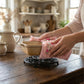Hands using pink floral lace pot holders to place a ceramic dish onto a black metal trivet on a wooden table.