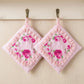 Two square pink pot holders with rose patterns and lace ruffles hanging from brass hooks against a light wood wall.
