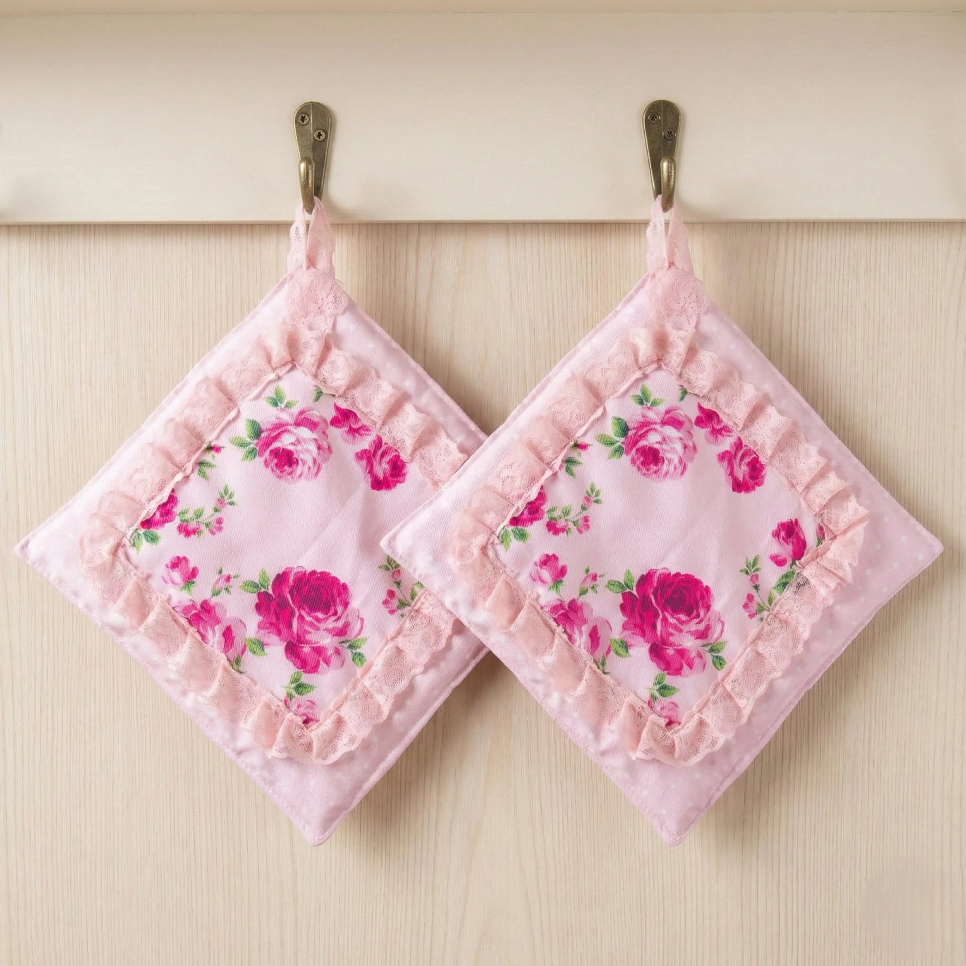 Two square pink pot holders with rose patterns and lace ruffles hanging from brass hooks against a light wood wall.