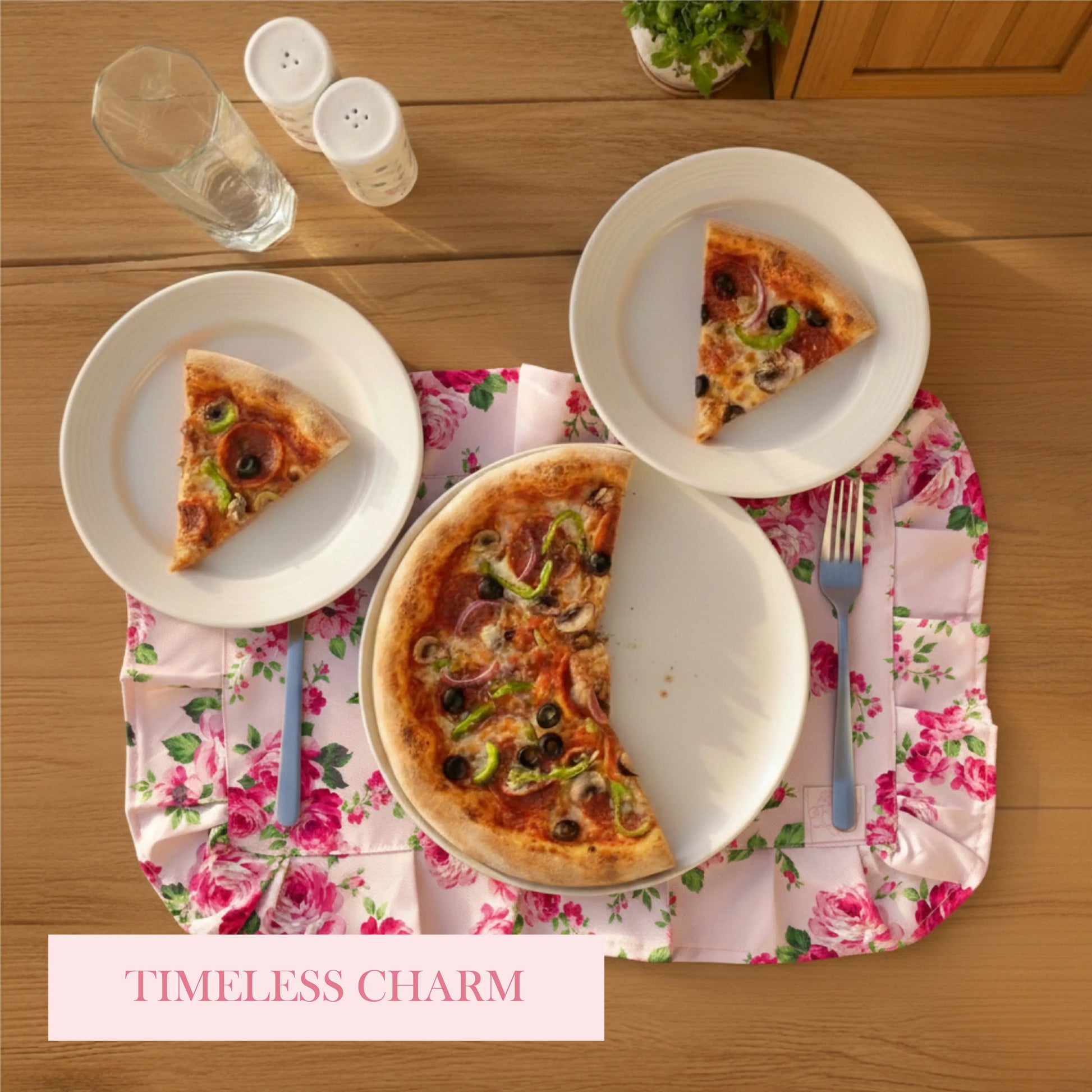 Pizza on a pink floral ruffled placemat with two side plates, water glass, and "TIMELESS CHARM" text on a wood table.