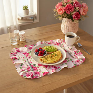 Breakfast plate on a pink floral ruffled placemat with a pink vase of flowers, coffee mug, and utensils on a wood table.