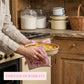 Hands using pink ruffled floral pot holders to hold a round baking dish in a rustic kitchen setting with jars.