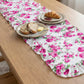 Top-down perspective of a pink floral ruffled table runner on a light wood surface, styled with breakfast plates featuring bread, butter, and a croissant with jam.