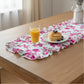 Pink floral ruffled table runner styled with a glass of orange juice and a stack of pancakes on a white plate over a natural wood table.