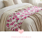 White ruffled runner with pink rose patterns laid across a neutral bed with beige pillows and blankets.