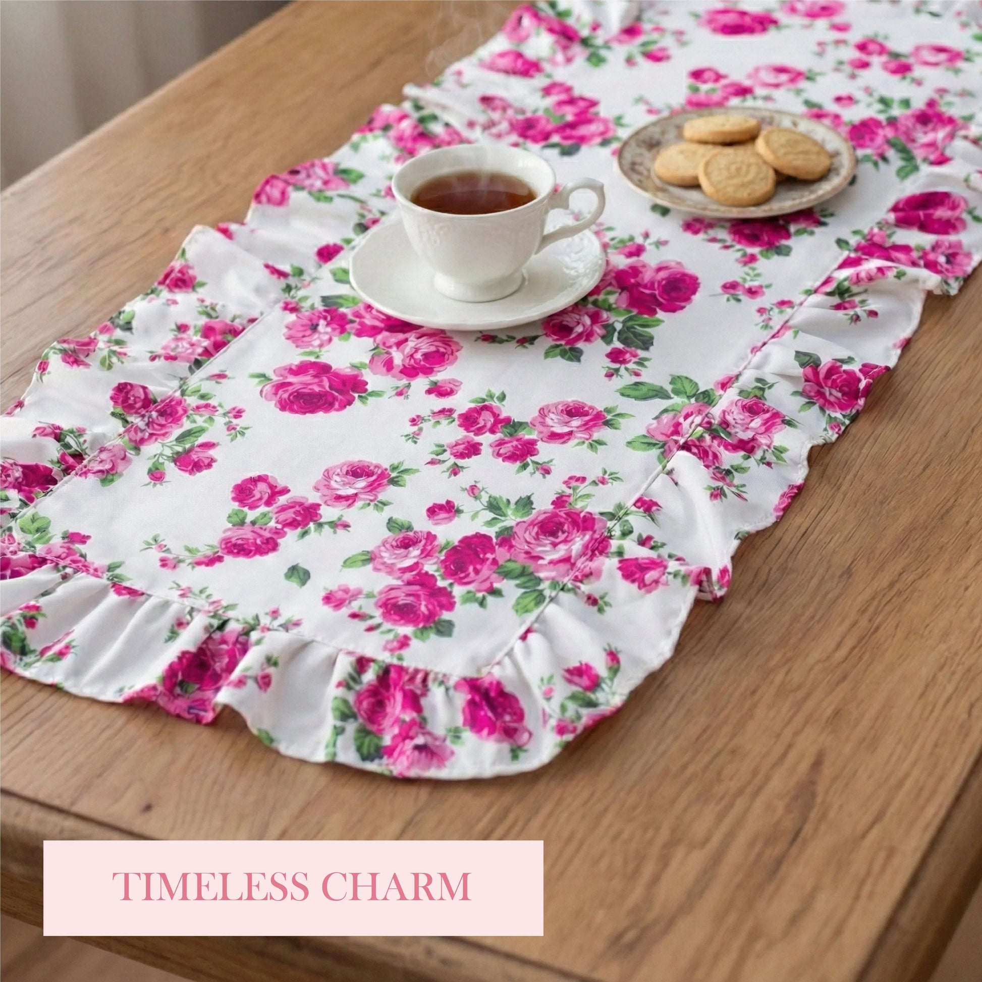 A white ruffled floral table runner with pink roses, a white teacup, and a plate of cookies on a wooden table.