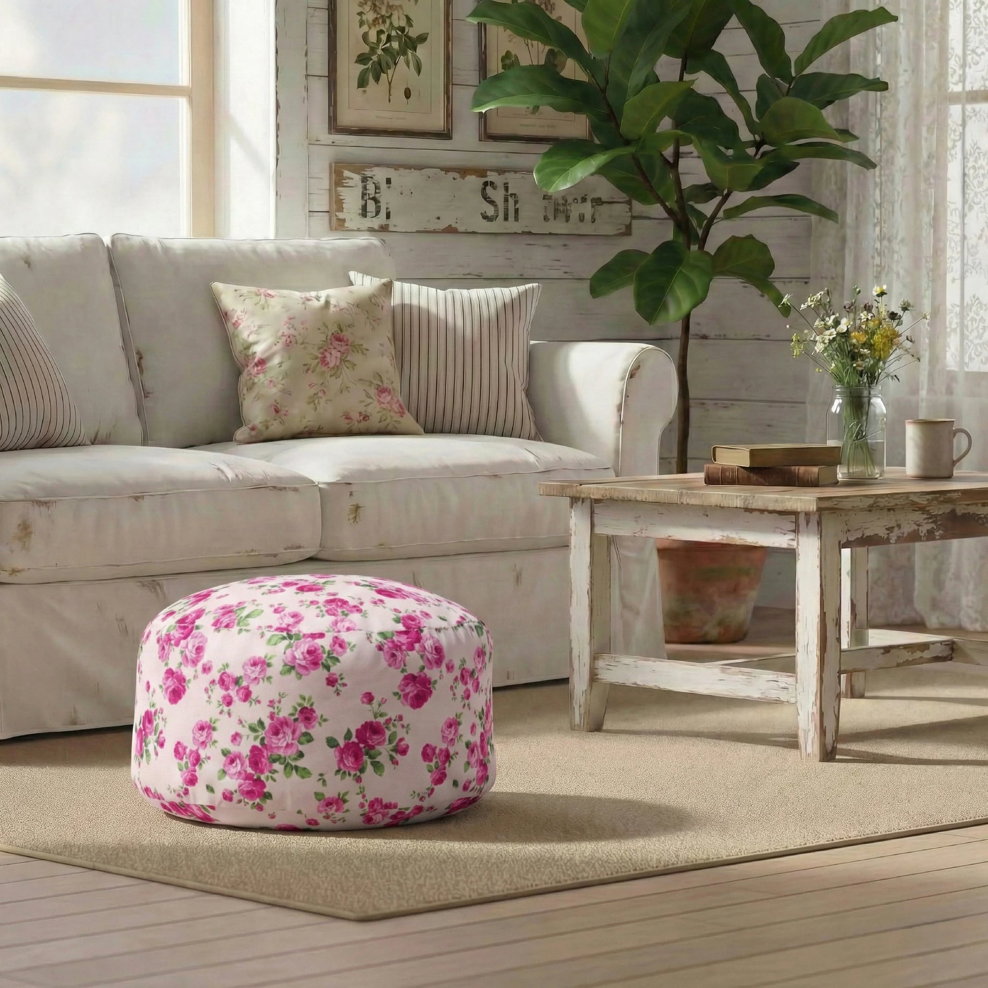 A pink rose fleur pouf on a beige rug in a farmhouse living room with a white slipcovered sofa, rustic wood coffee table, and large indoor plant.