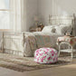 A pink rose fleur pouf centered on a textured rug at the foot of a distressed white wooden bed in a bright, vintage-style bedroom.