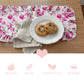 Chocolate chip cookies on a white plate over a pink floral ruffled table runner with product feature icons below.