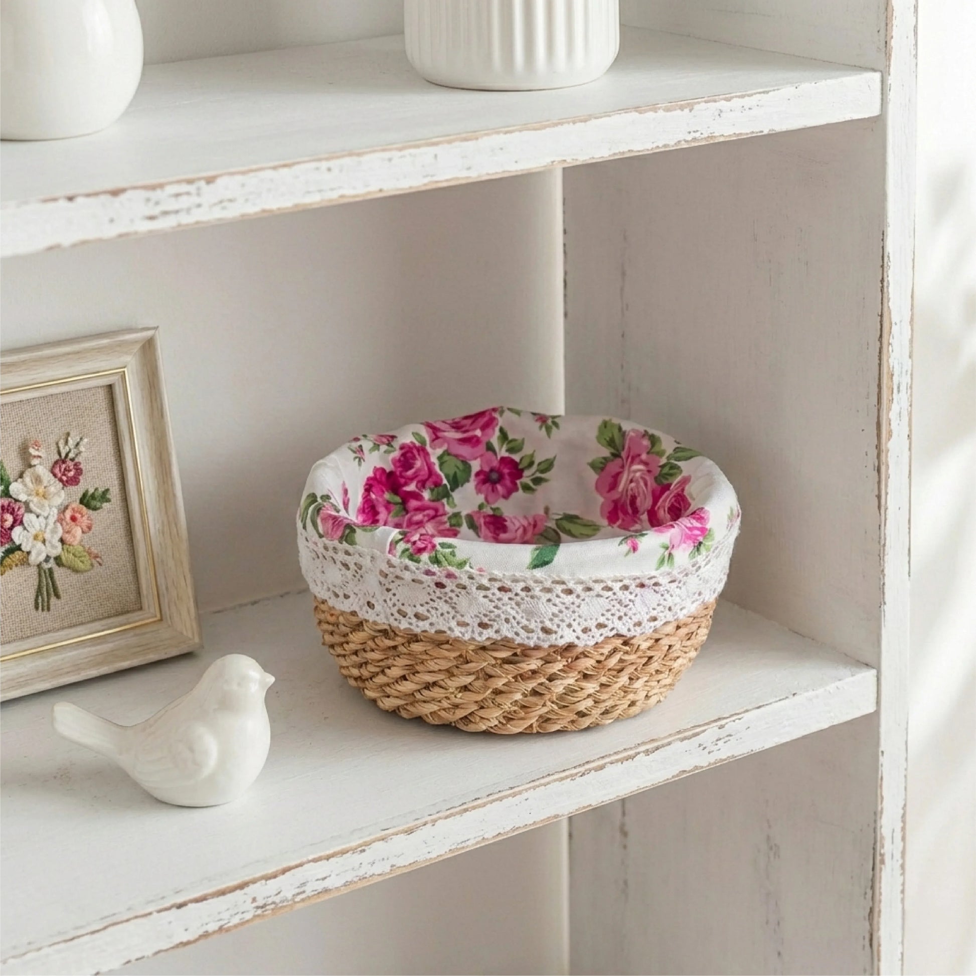 A round handwoven halfa grass basket with a pink rose fabric liner and white lace trim, displayed on a distressed white wooden shelf next to a small bird figurine and a floral picture frame.