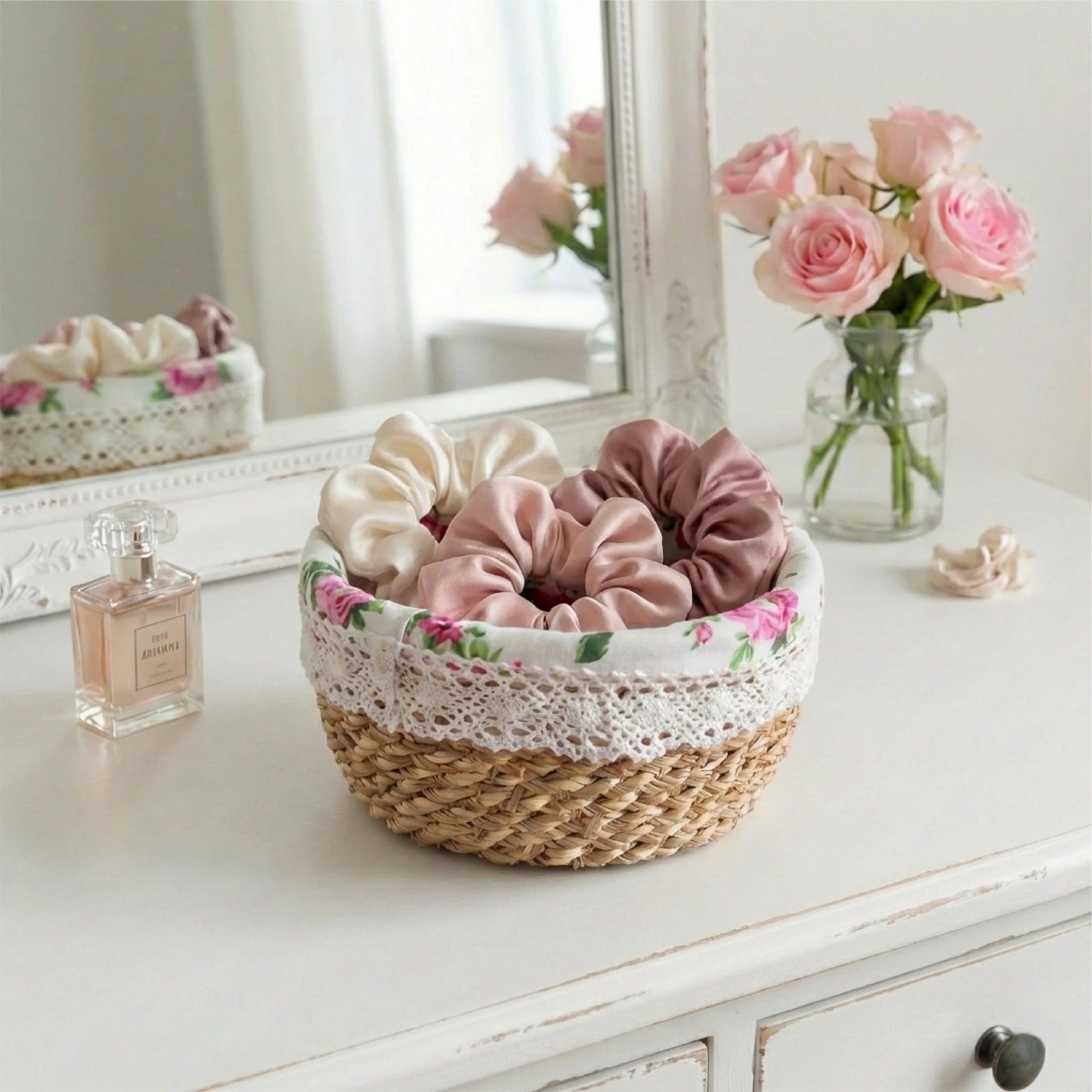 Round fleur basket holding silk scrunchies on a white vanity next to a perfume bottle and pink roses.