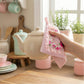 A hand holding a cream teapot with a pink ruffled floral pot holder in a bright kitchen with plants.