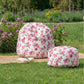 A floral bean bag and matching fleur pouf with a pink rose pattern set on a natural jute runner in a lush green garden surrounded by flowers.
