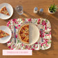Top-down view of pizza slices on white plates, a floral ruffled placemat, and a water glass on a wooden table.