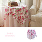Floral tablecloth on a round table with books and a vase, next to a beige armchair.