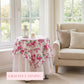 Floral tablecloth on a round table in a bright room with a couch and windows.