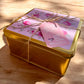Decorative gold box with floral lid and pink tag on a wooden surface