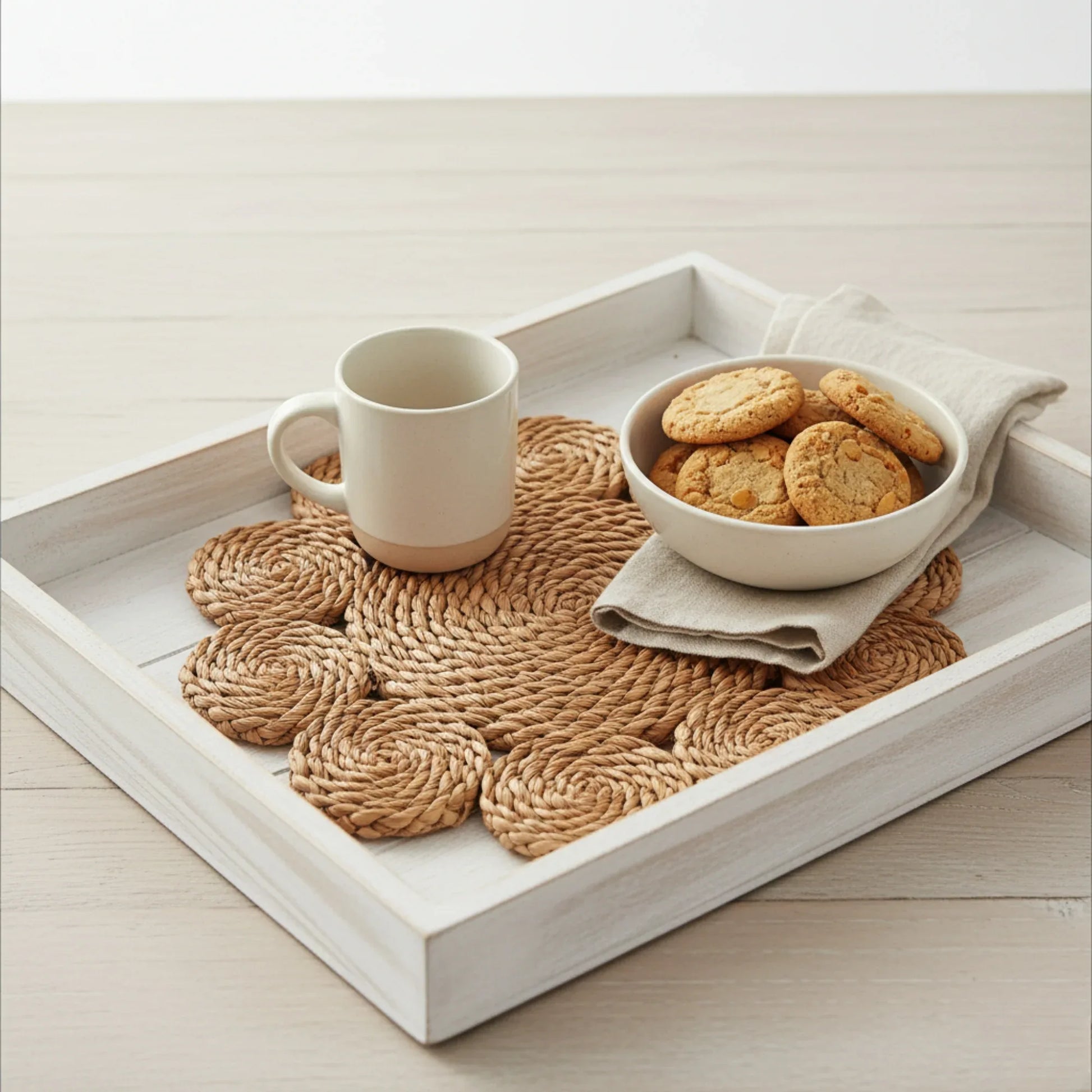 White tray with woven placemat, mug, bowl of cookies, and napkin on a light wooden surface.