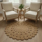 Round jute rug in front of two beige armchairs with a small wooden table and plant in a bright room.
