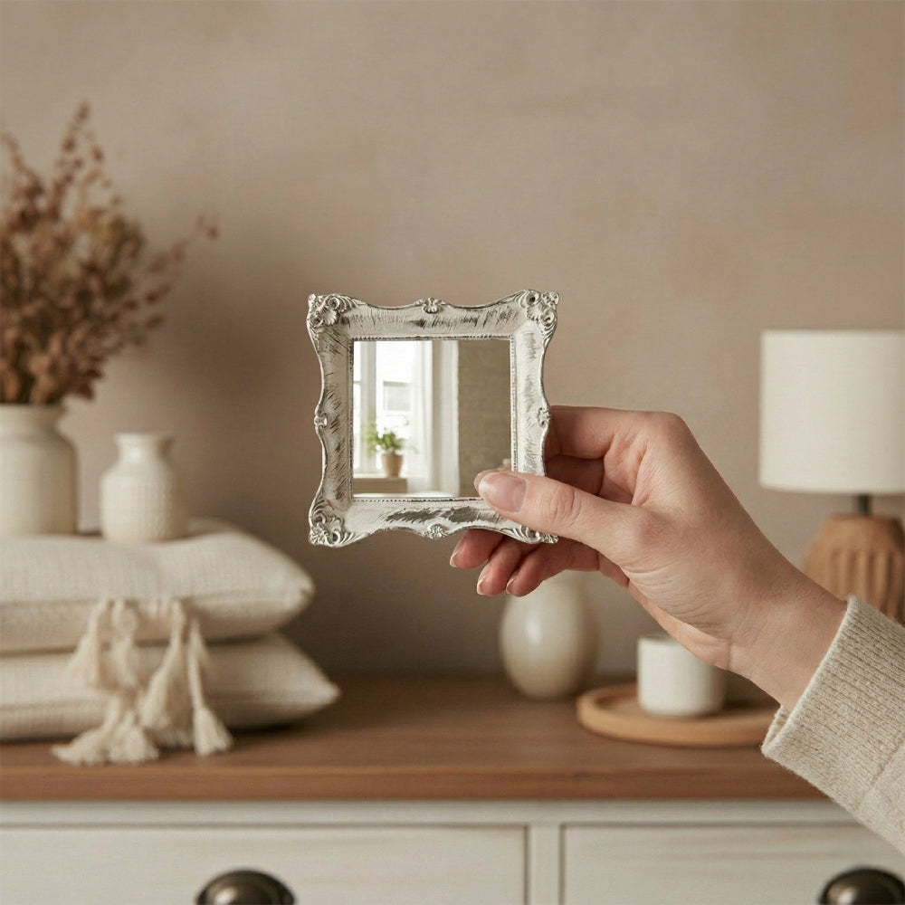 A hand holding a small antique white ornate mirror in front of a dresser with pillows, a lamp, and a vase.