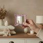 A hand holding a small pink ornate mirror in front of a dresser with neutral pillows, a lamp, and dried plants.