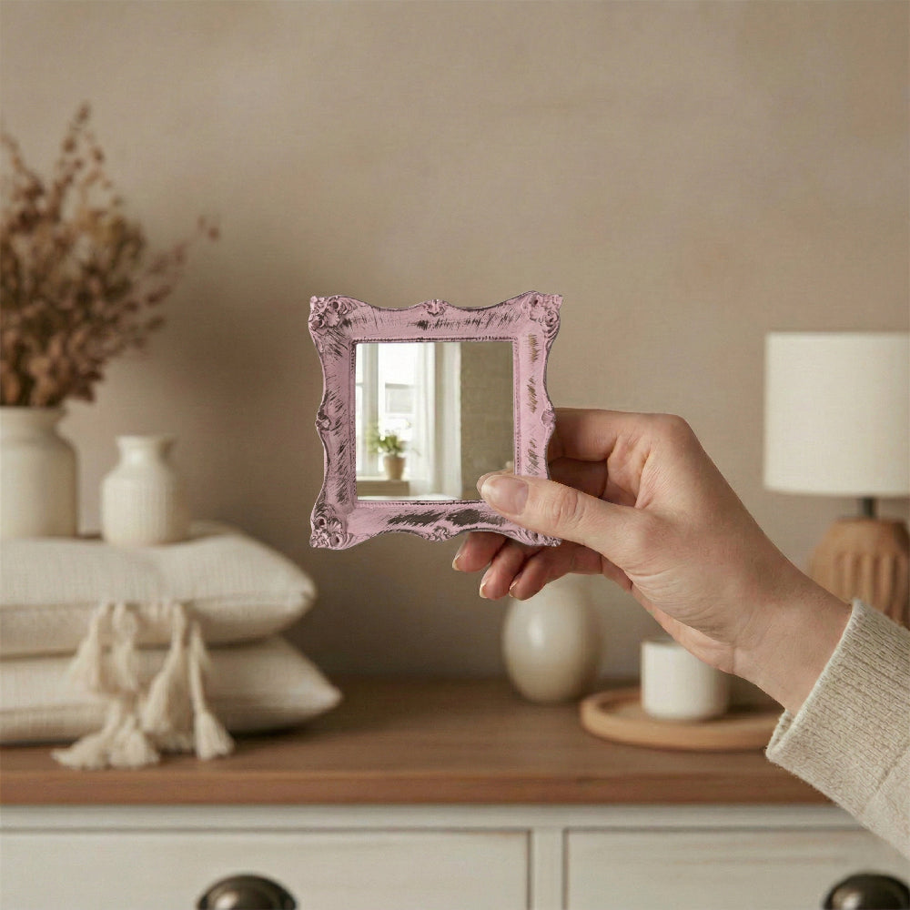 A hand holding a small pink ornate mirror in front of a dresser with neutral pillows, a lamp, and dried plants.
