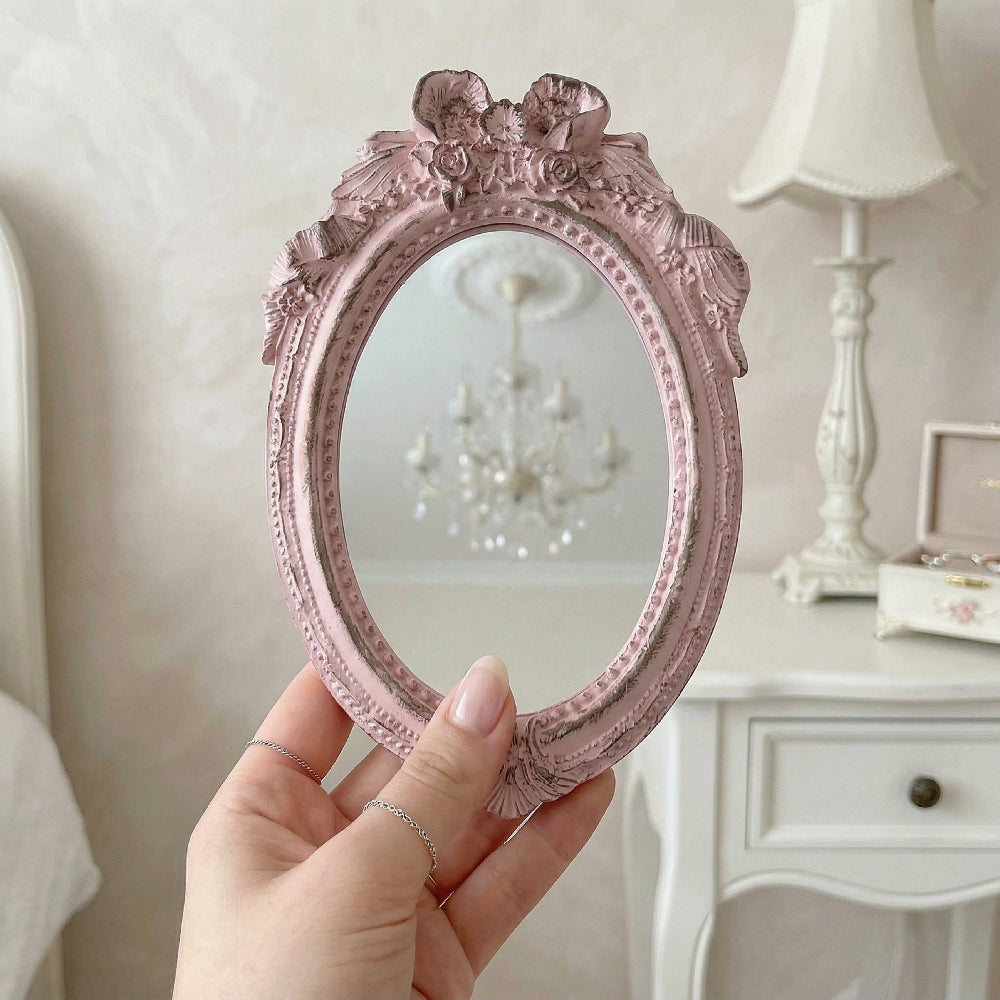 A hand holding a small pink ornate oval mirror with floral details, reflecting a chandelier in a bedroom setting.