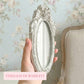 A hand holding a small, ornate oval mirror with an antique white distressed frame against floral wallpaper.