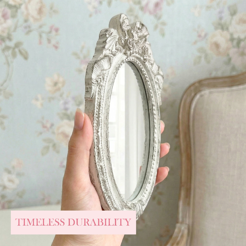 A hand holding a small, ornate oval mirror with an antique white distressed frame against floral wallpaper.