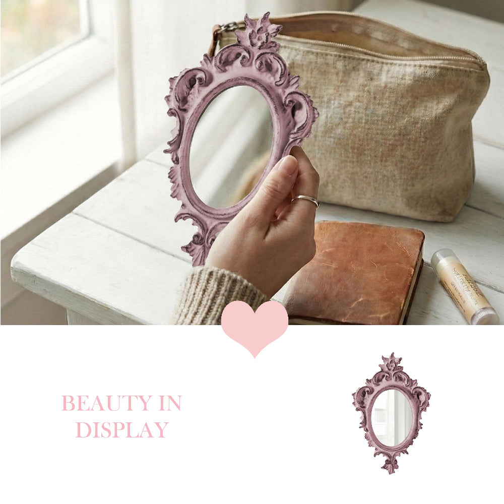 A hand holding a dusty pink ornate mirror near a linen bag and leather journal on a white table with pink text.